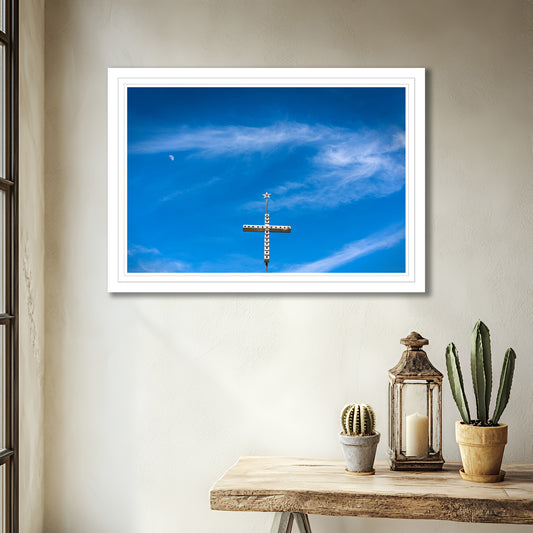 White frame shown in room. Framed photo of a cross made from birdhouses, crowned with a star, under a blue Texas sky with light clouds and the moon visible.